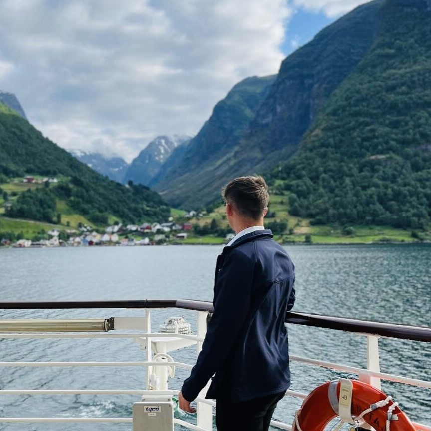 A person stands on a boat deck, part of the Carnival Corporation cadet programme, gazing at a scenic fjord with green mountains, a village, and calm water under a cloudy sky. A lifebuoy hangs on the railing nearby.