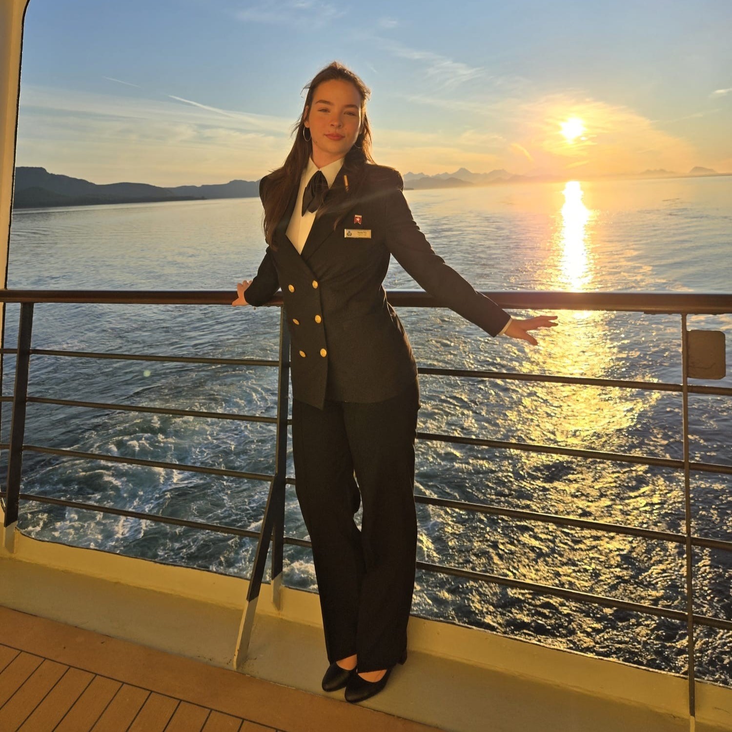 A woman in a navy uniform, part of the Carnival Corporation cadet programme, stands on a ship’s deck at sunset, holding the rail as golden sunlight illuminates the ocean and distant mountains.