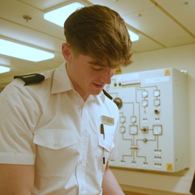 A young man in a white uniform with shoulder epaulettes and a name badge stands indoors, looking down. Behind him is a control panel with buttons and diagrams, hinting at his role in the Carnival Corporation cadet programme.