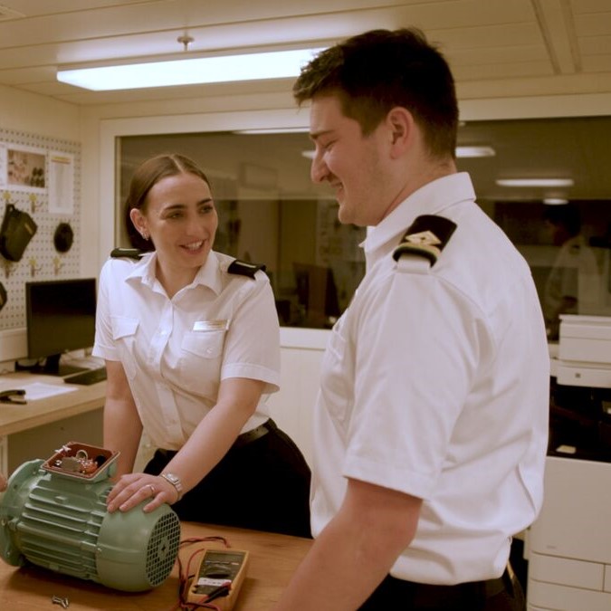 Two people in uniform stand at a workbench, smiling at each other. One person rests a hand on a green motor with tools nearby, suggesting they may be part of the Carnival Corporation cadet programme in a technical or engineering workspace.
