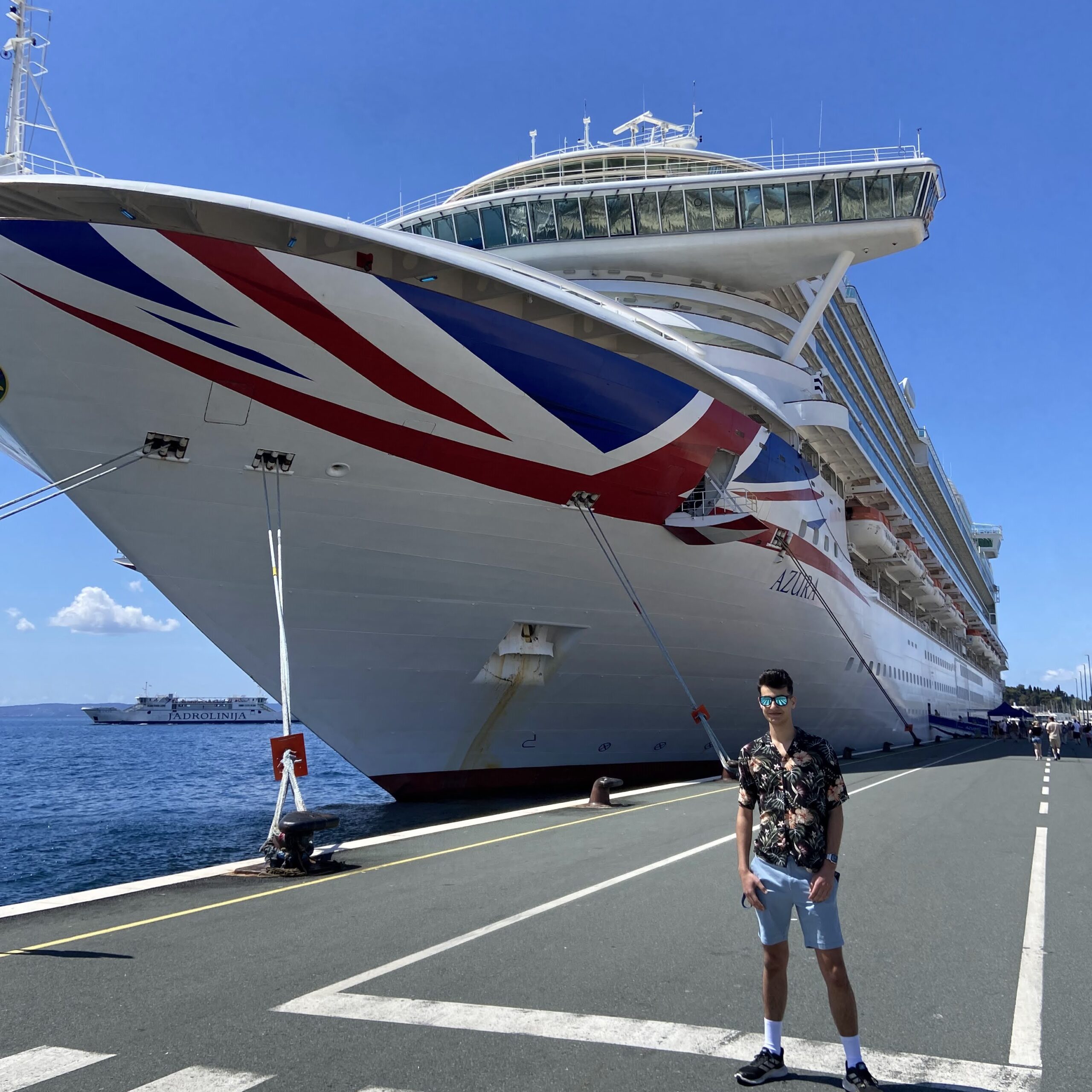 A man wearing sunglasses, a floral shirt, and shorts stands on a pier in front of a large cruise ship with a red, white, and blue design—showcasing the Carnival Corporation cadet programme—under a clear blue sky.