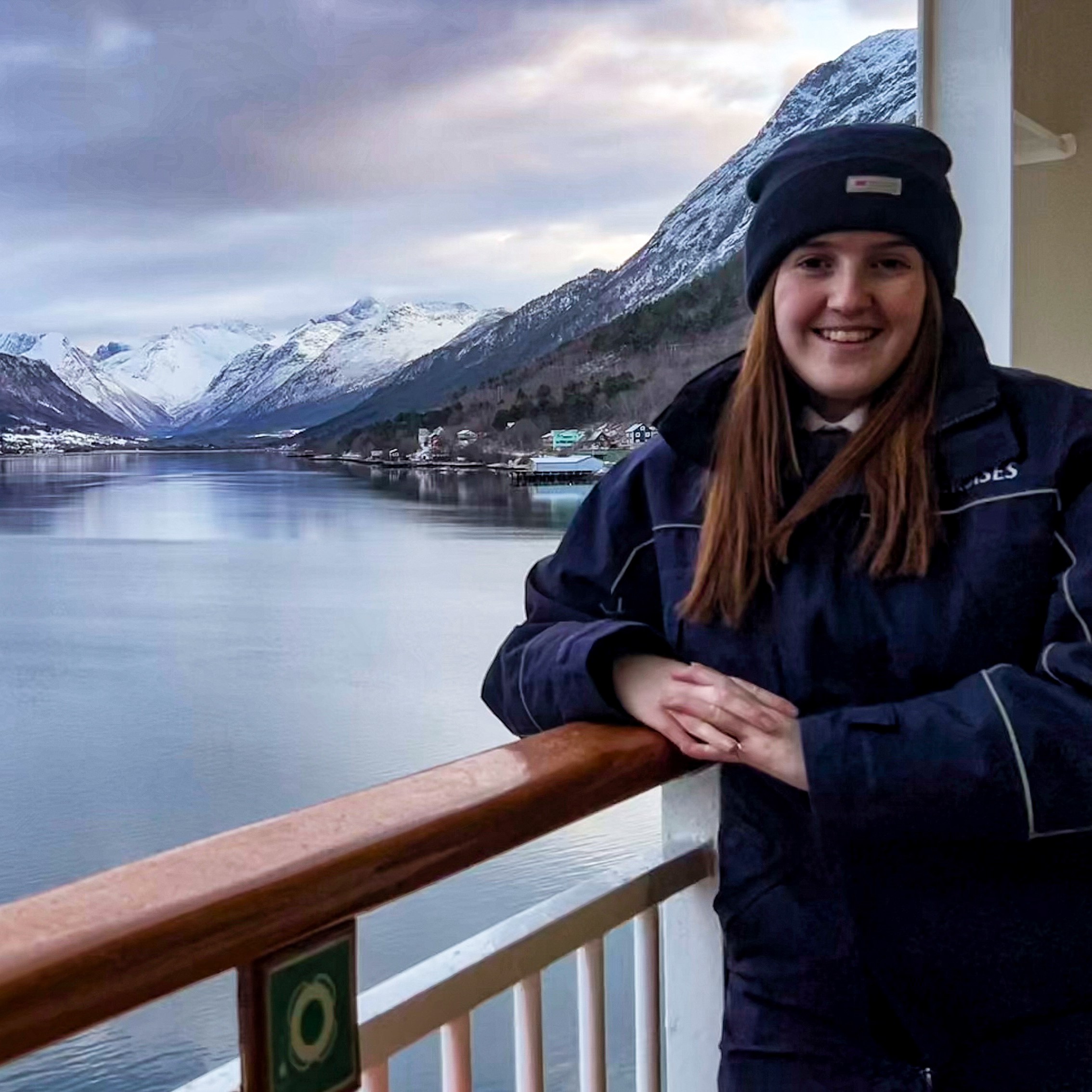 A woman in a winter coat and hat, part of the Carnival Corporation cadet programme, smiles whilst standing on a balcony overlooking a calm fjord surrounded by snow-covered mountains and scattered houses under a cloudy sky.
