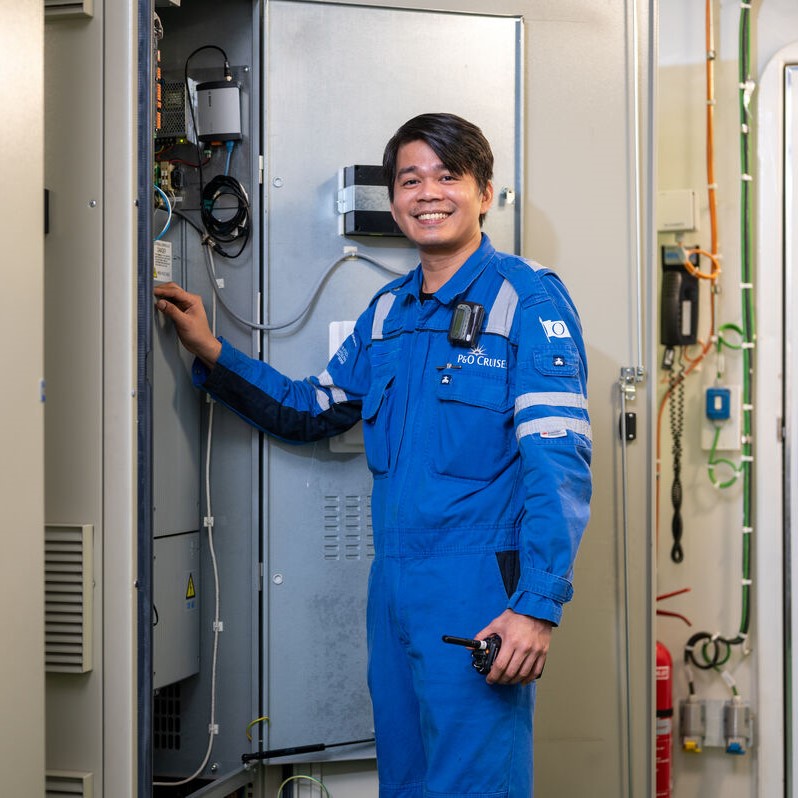 A smiling engineer in a blue boiler suit stands next to an open electrical panel, holding a walkie-talkie, showcasing the dynamic work environment offered by P&O Cruises technical careers in a brightly lit industrial setting.