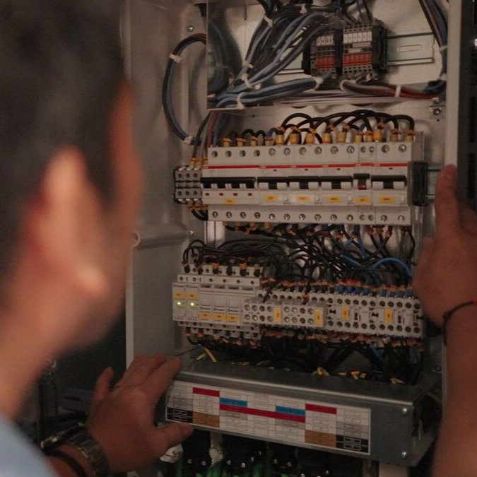 A person examines an open electrical control panel filled with wires, switches, and circuit breakers—tasks typical in P&O Cruises technical careers. The individual's hands are on the panel, with wiring and components clearly visible inside.