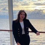 A woman in a dark blazer and white shirt stands by a railing on a boat or ship, with the sea and a cloudy sunset sky in the background.