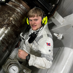 A person in a white protective suit and yellow ear defenders is working with machinery. They are looking up and smiling at the camera whilst holding a valve or lever. A round pressure gauge is visible nearby.