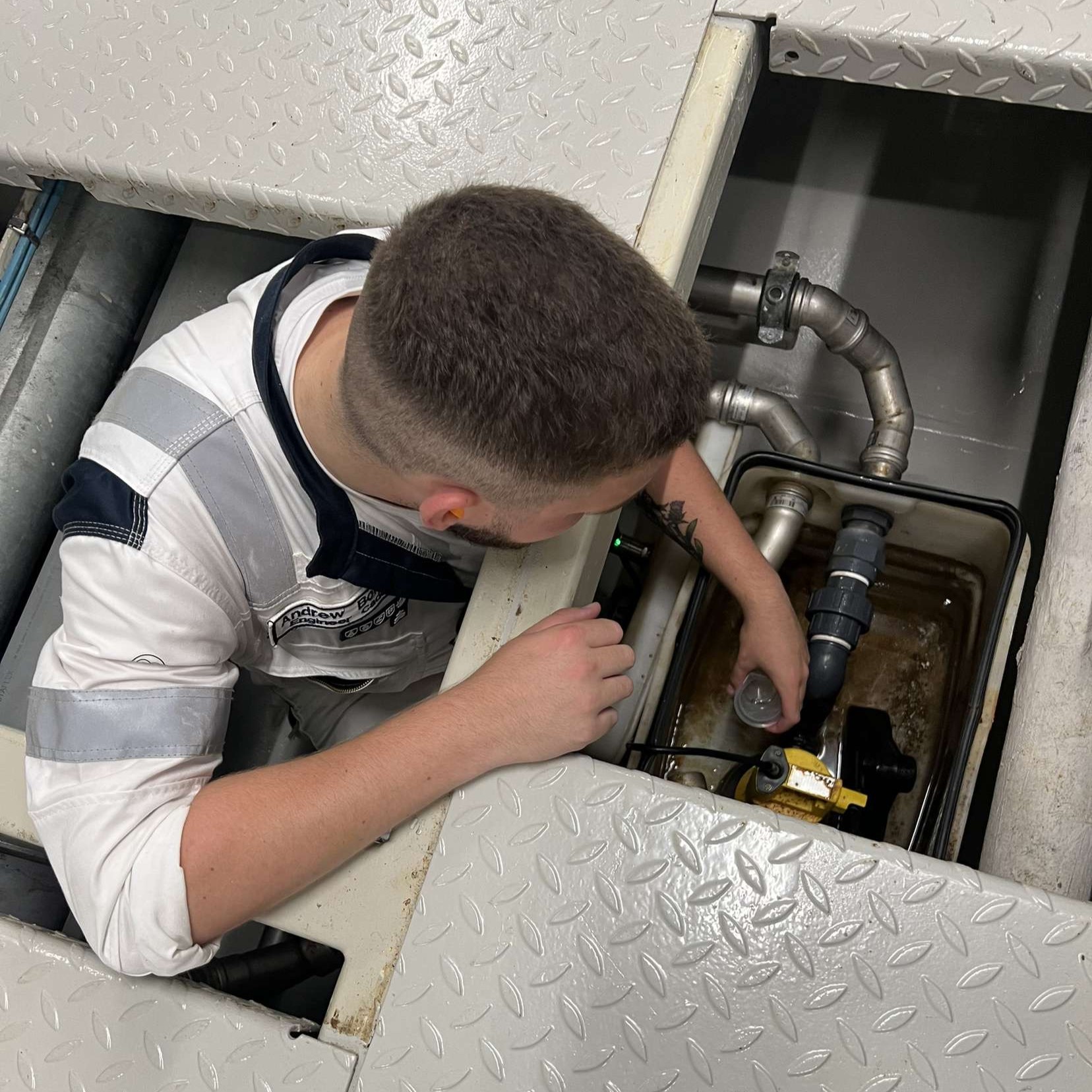 A person wearing a striped shirt and dungarees, possibly part of the Carnival Corporation cadet programme, works inside a metal compartment inspecting or adjusting pipes and valves in an industrial area with a chequer-plate floor and plumbing components.