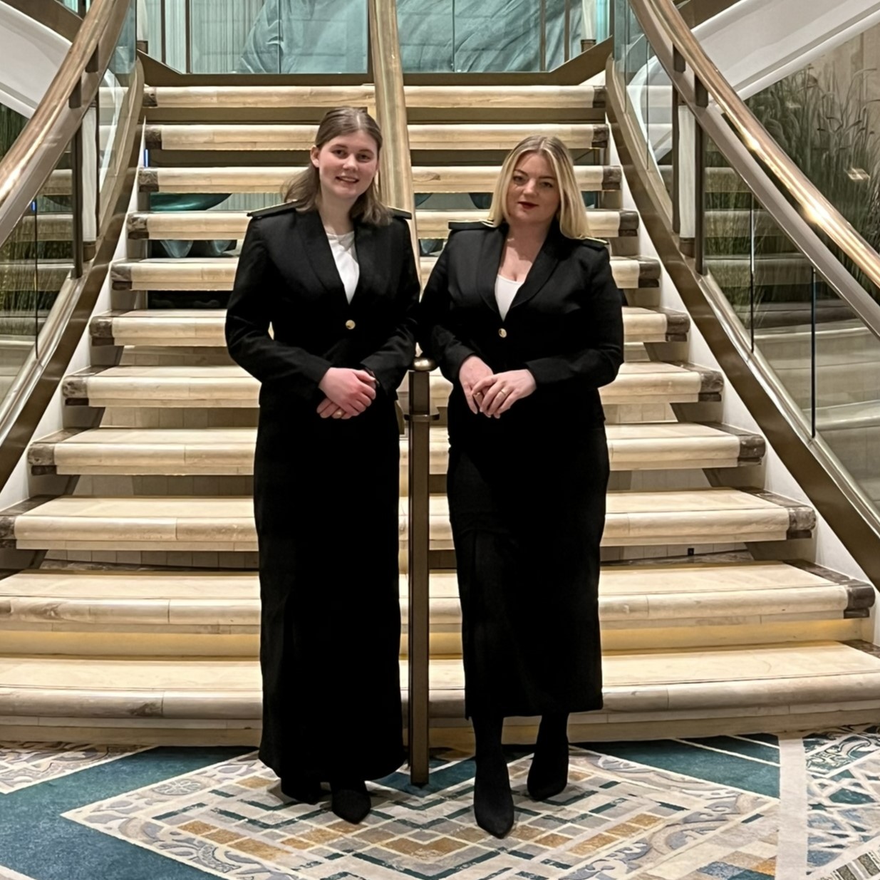 Two women, part of the Carnival Corporation cadet programme, stand side by side in front of an elegant staircase. Both wear long black blazers and skirts with white shirts, set against polished railings and a patterned floor.