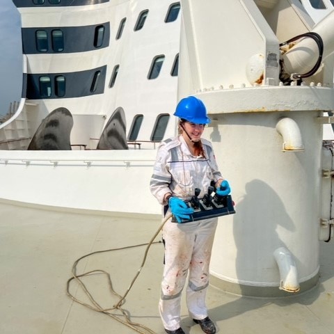 A person in a blue hard hat, gloves, and white coverall stands on a ship deck holding equipment—possibly participating in the Carnival Corporation cadet programme—with anchor mechanisms and part of the ship's superstructure visible behind.