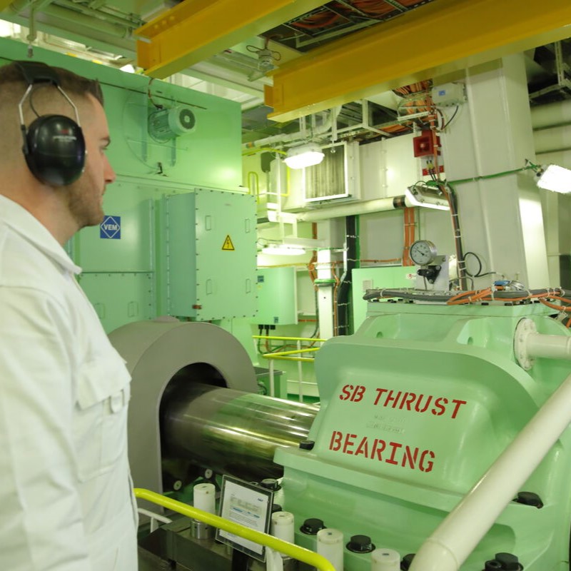 A man in white overalls and headphones observes a large industrial machine labelled SB THRUST BEARING inside a brightly lit, modern facility—an environment reflecting the expertise found in P&O Cruises technical careers.