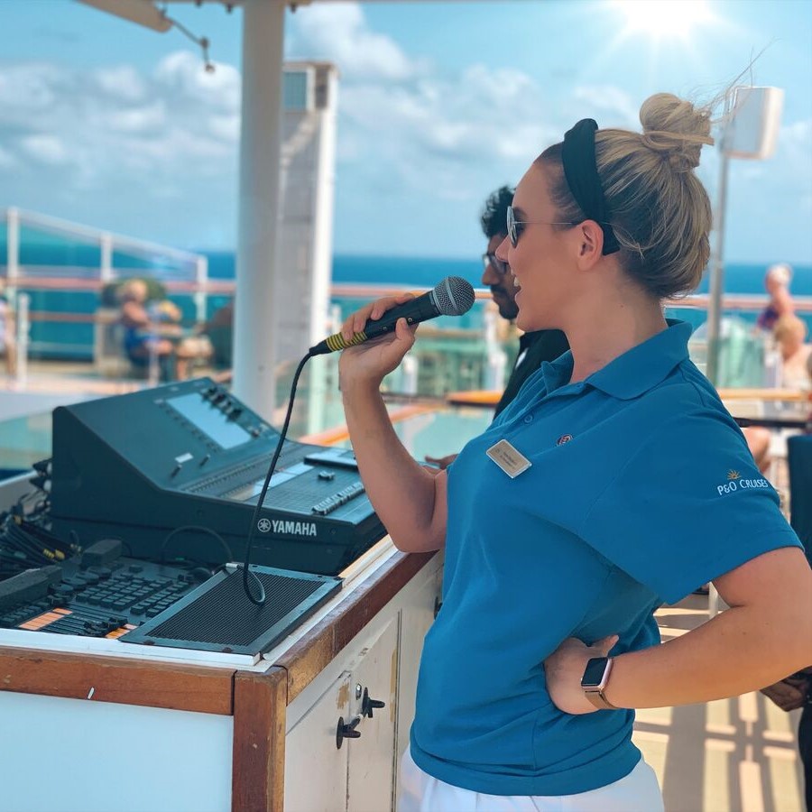 A woman in a blue polo shirt holds a microphone and speaks beside sound equipment on a sunny outdoor deck, with the sea and blue sky in the background.