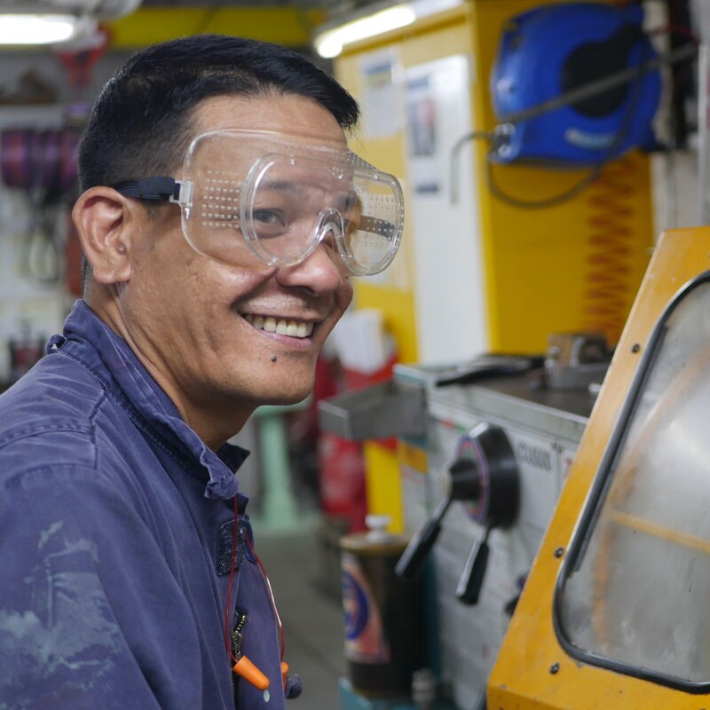 A man wearing safety goggles and a blue work shirt smiles whilst standing next to industrial machinery in a workshop, highlighting the hands-on expertise found in P&O Cruises technical careers.