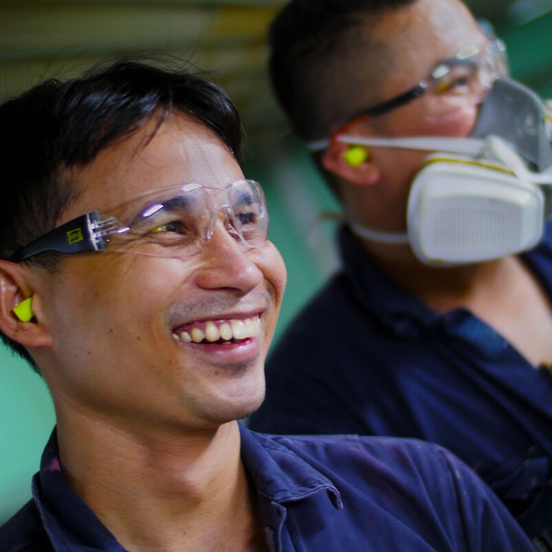 Two men in an industrial workshop wear safety goggles, earplugs, and dark shirts. One smiles while the other wears a respirator mask—showcasing the safety focus of P&O Cruises technical careers.