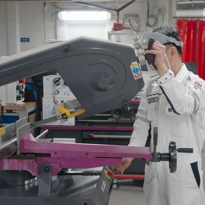 A person in a white industrial uniform operates a large bandsaw machine in a workshop, demonstrating the precision and expertise found in P&O Cruises technical careers. The workspace is equipped with various tools and equipment.