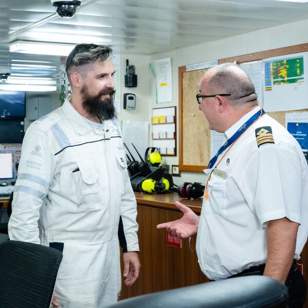 Two men in uniform, one in white overalls and the other in a white shirt with captain’s stripes, discuss P&O Cruises technical careers in an office lined with safety equipment and noticeboards.