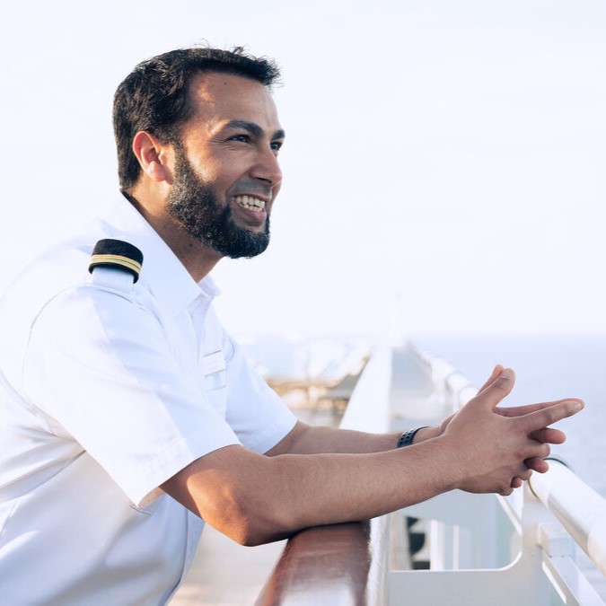 A smiling man in a white officer’s uniform stands on the deck of a ship, leaning on the rail and looking out over the sea on a bright, clear day.