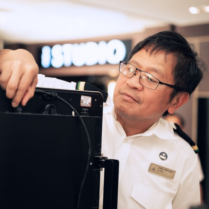 A man wearing glasses and a white uniform shirt with a name badge adjusts electronic equipment indoors, with a blurred STUDIO sign in the background.