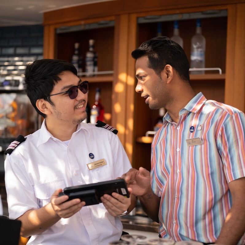 Two men in uniform stand behind a bar, one in a white shirt holding a tablet and the other in a striped shirt, engaged in conversation with bottles on shelves in the background.