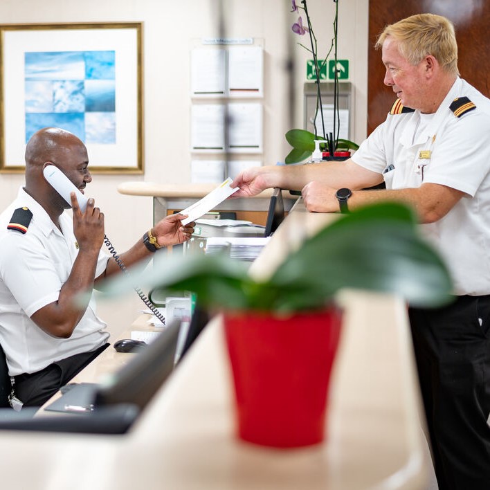 Two uniformed men work at a reception desk; one is seated and talking on the phone whilst taking a document from the other, who is standing. A potted plant is in the foreground, partially out of focus.
