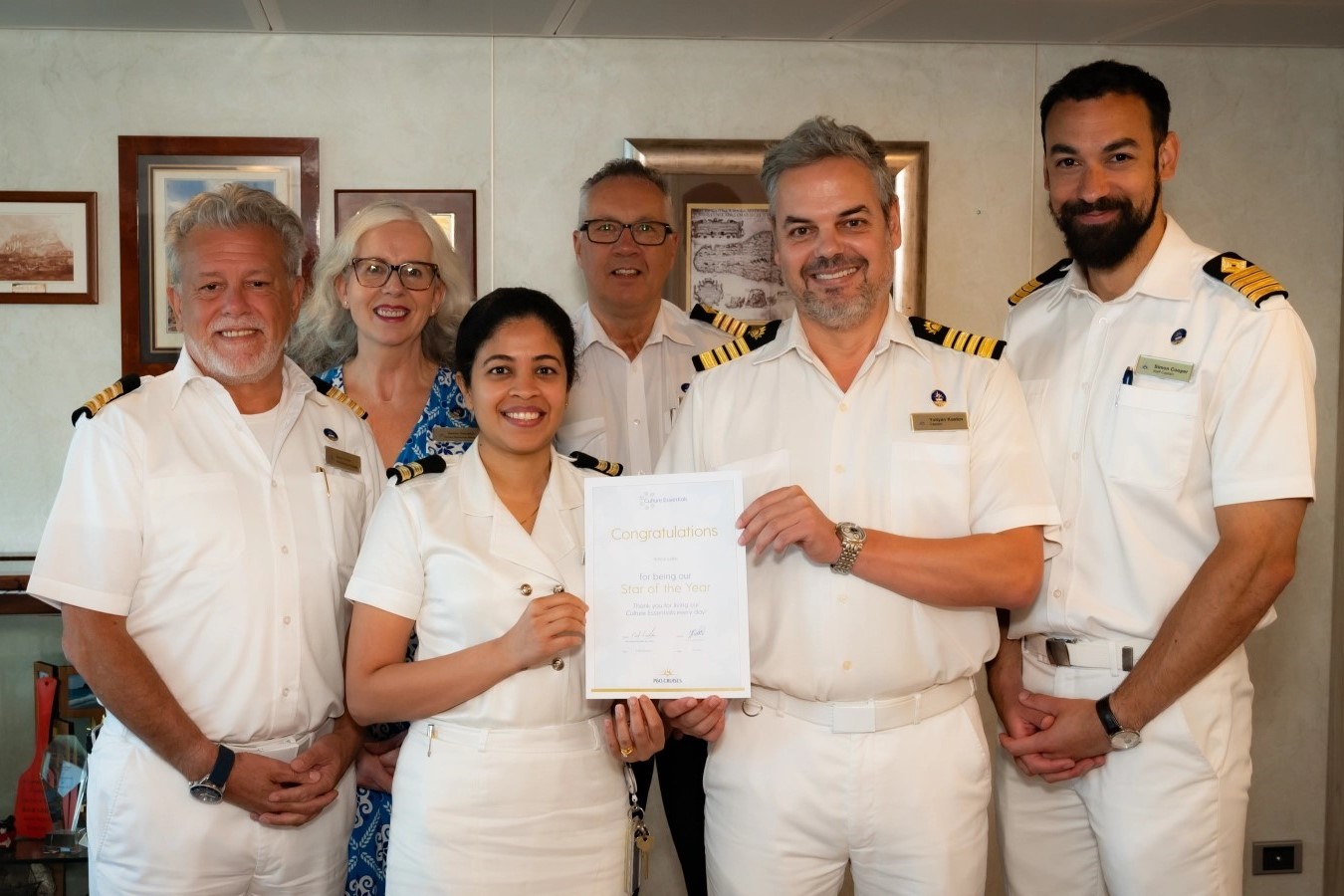 Six smiling cruise ship staff in uniforms stand indoors. The woman at front centre holds a certificate that reads “Congratulations” with her colleagues gathered around her, celebrating her achievement. Framed photos hang on the wall behind them.
