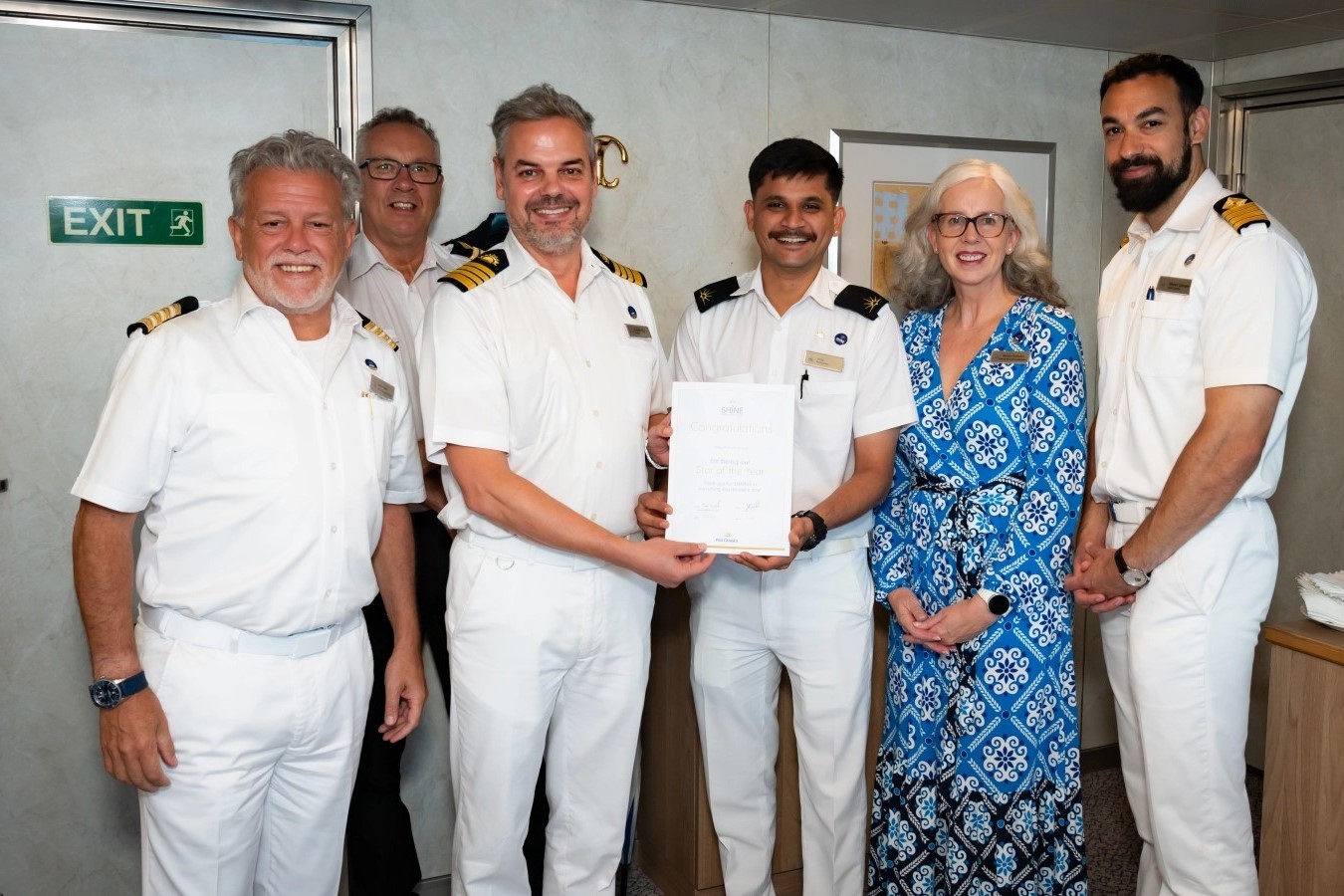 Six people, including four men in white uniforms, one man in a suit, and one woman in a blue patterned dress, stand together indoors. Two men in uniforms hold a certificate and all are smiling at the camera.