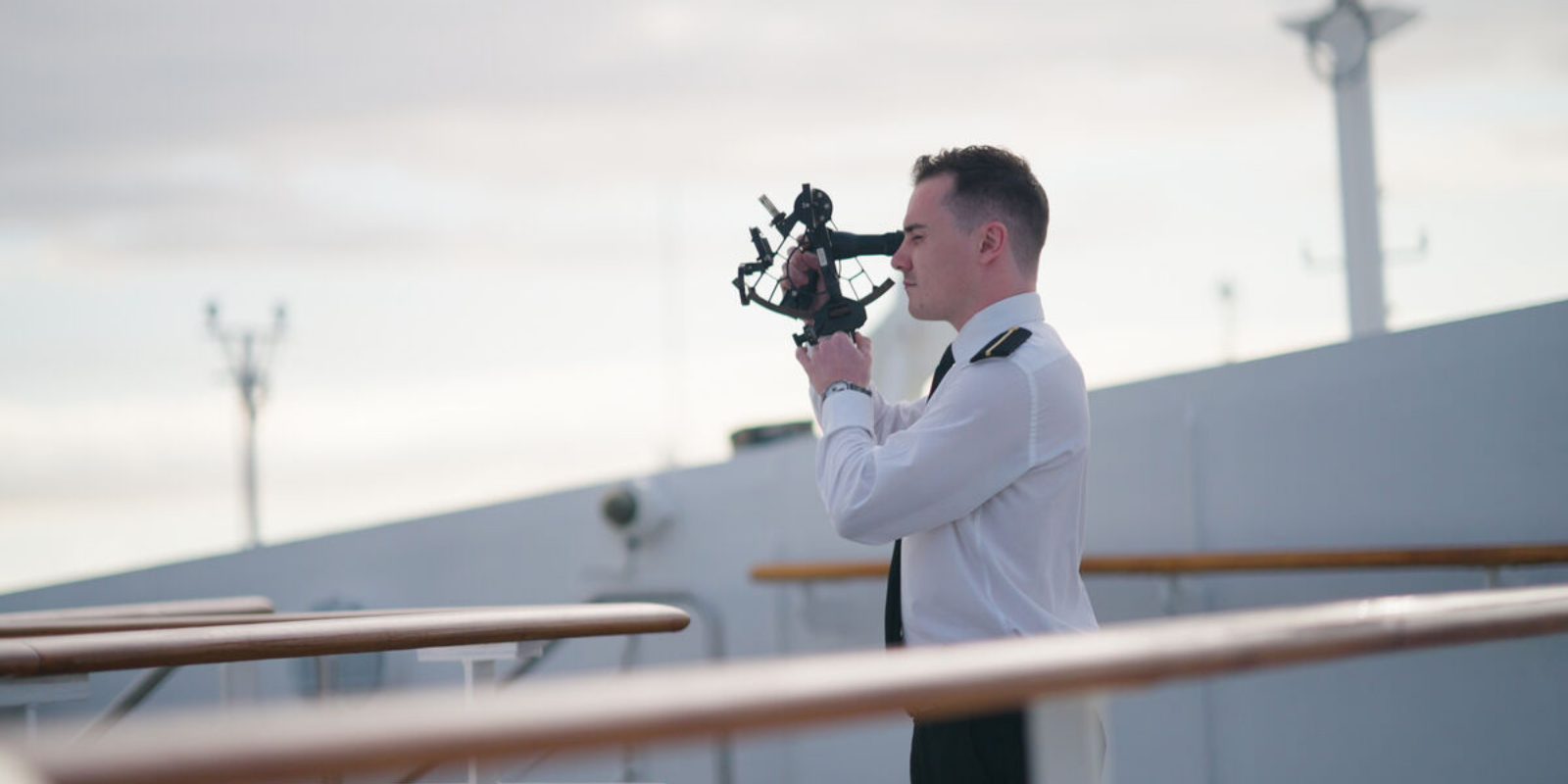 A person in a white uniform stands on a ship’s deck using a sextant to navigate—part of the Carnival Corporation cadet programme—with rails and equipment visible under a cloudy sky.