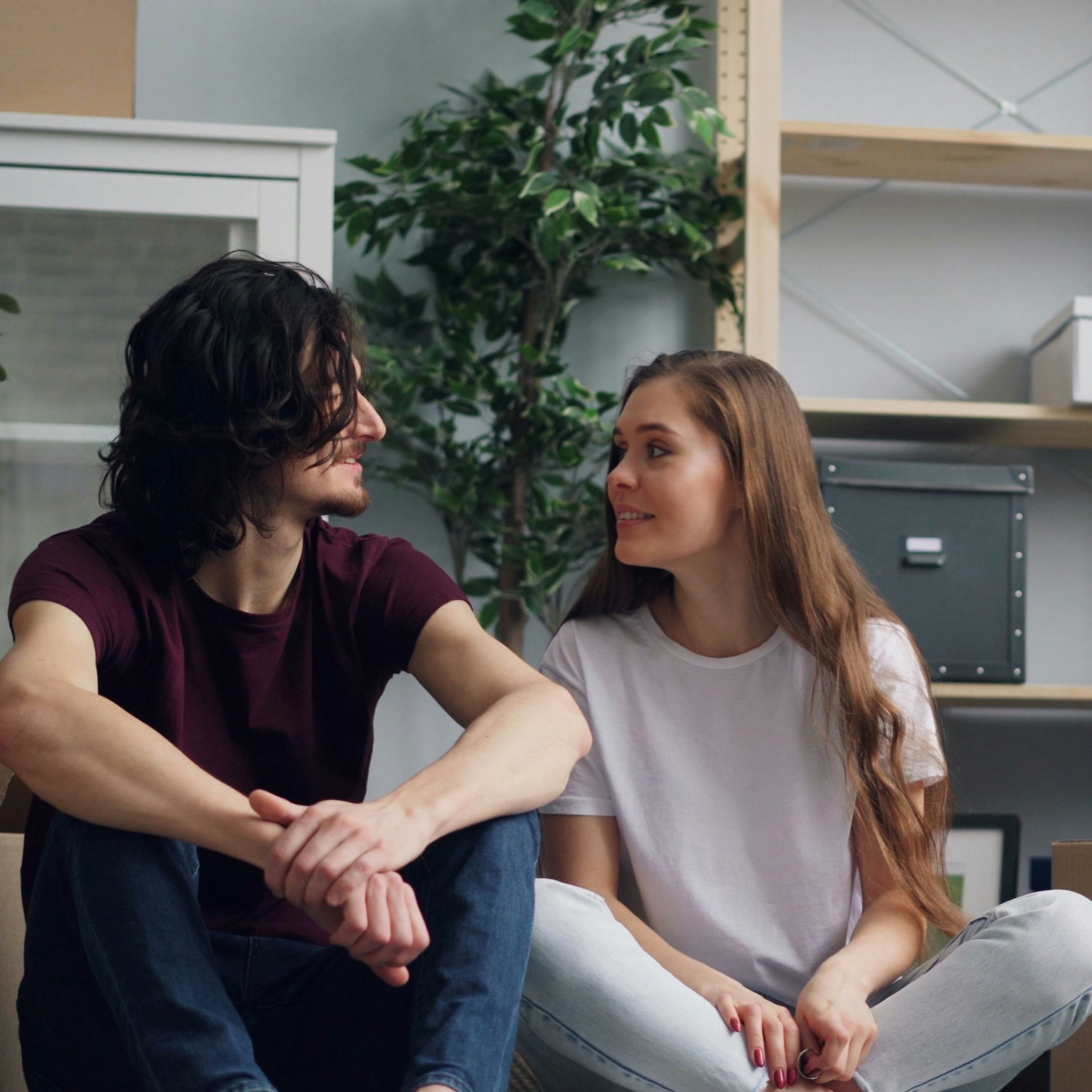 A man and a woman sit cross-legged on the floor indoors, smiling at each other—perhaps discussing their experiences in the Carnival Corporation cadet programme. Shelves, a potted plant, and storage boxes are visible in the background.