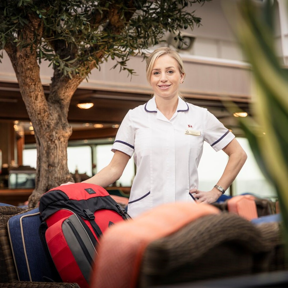 A woman in a white uniform stands smiling beside a red rucksack on a chair, in an outdoor lounge area with greenery and wicker furniture in the background.