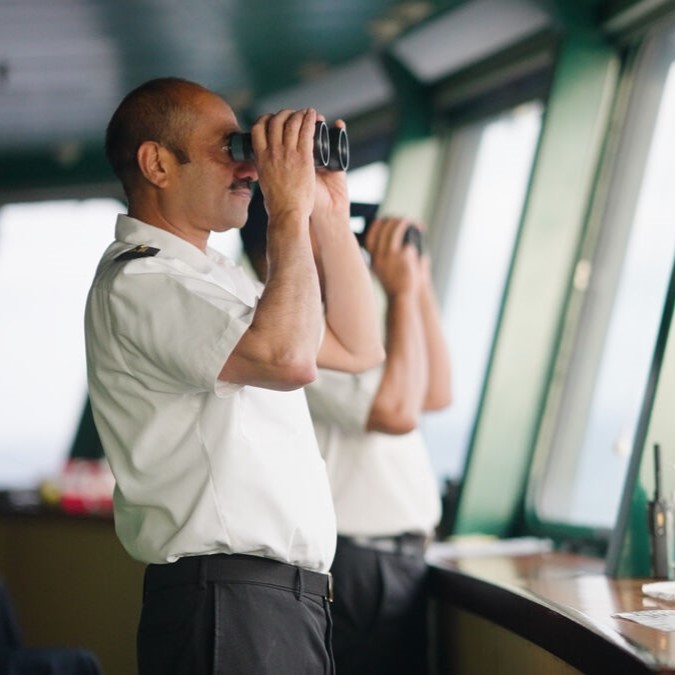 Two uniformed ship officers stand on a vessel’s bridge, each scanning the sea ahead through binoculars. A radio and papers rest on the console—offering a glimpse into P&O Cruises deck careers in action.