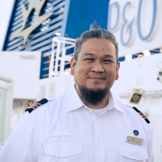 A cruise ship crew member in a white uniform with shoulder epaulettes and a name badge stands smiling on deck. The P&O Cruises logo and ship structure are visible in the background.