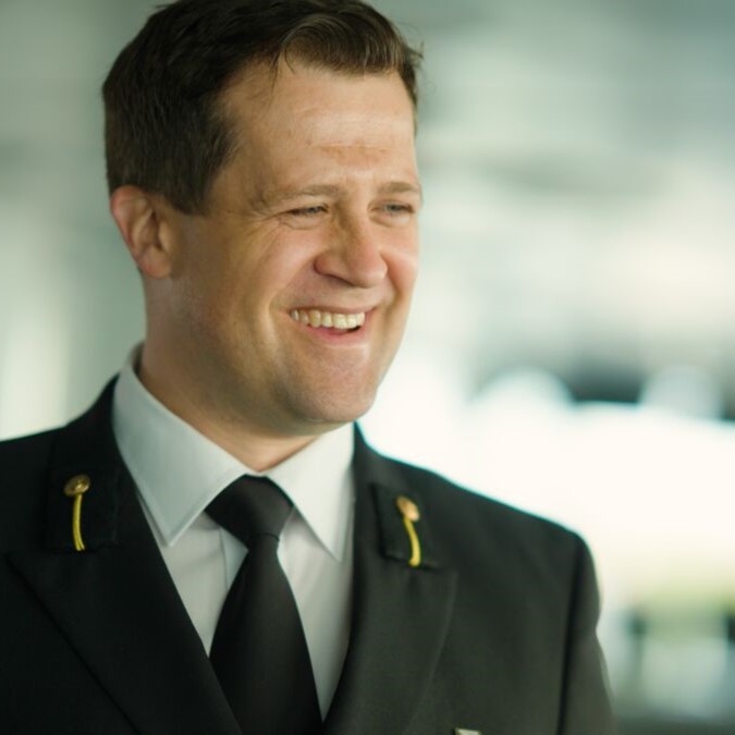 A man in a black uniform jacket with gold pins, part of the Carnival Corporation cadet programme, smiles and looks to the side. The background is blurred.