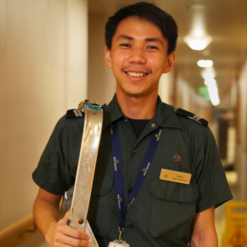 A smiling young man in a green uniform with a name tag and lanyard stands in a brightly lit corridor, holding a metallic object over his shoulder—a glimpse into P&O Cruises technical careers.