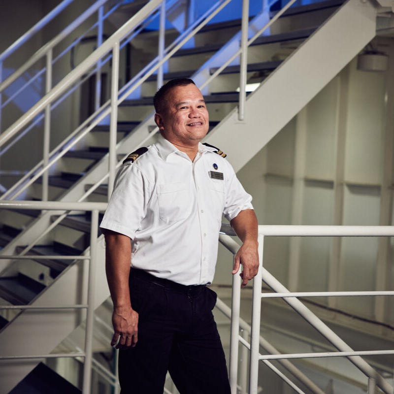 A man in a white uniform shirt and black trousers stands smiling by a staircase indoors. His shoulder epaulettes suggest he is part of the crew, possibly working in one of P&O Cruises technical careers.