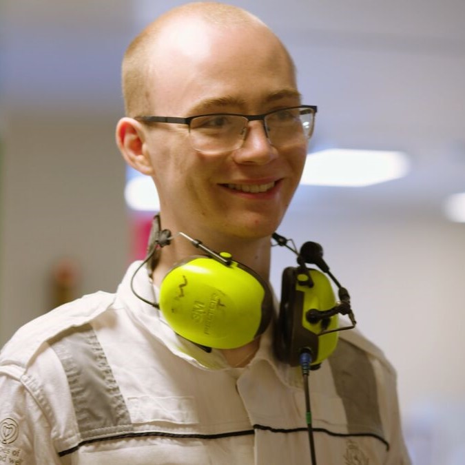 A smiling person with glasses and a short haircut wears a white shirt and has bright yellow ear defenders resting around their neck, ready for hands-on training in the Carnival Corporation cadet programme. The background is softly blurred.