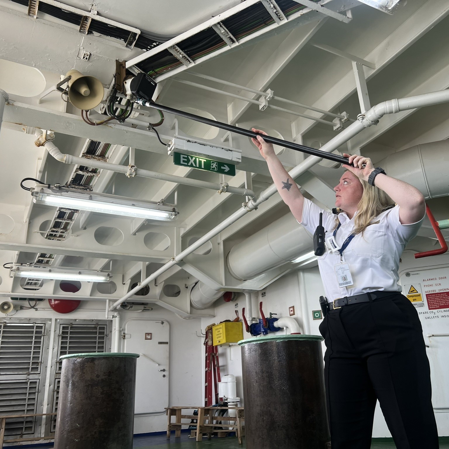 A uniformed woman, part of the Carnival Corporation cadet programme, stands indoors on a ship, using a long pole to clean or inspect an overhead area near a loudspeaker and pipes. There is an EXIT sign, emergency equipment, and safety signs visible in the background.