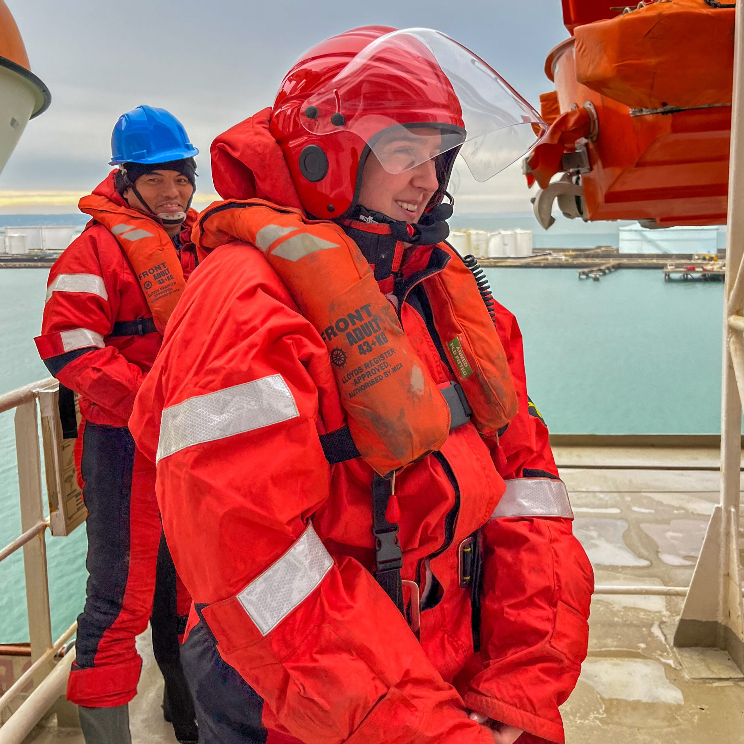 Two people in red survival suits and helmets stand on a ship’s deck by the water, likely participating in the Carnival Corporation cadet programme. The person in front wears a clear face visor and life jacket; city buildings appear in the distance.