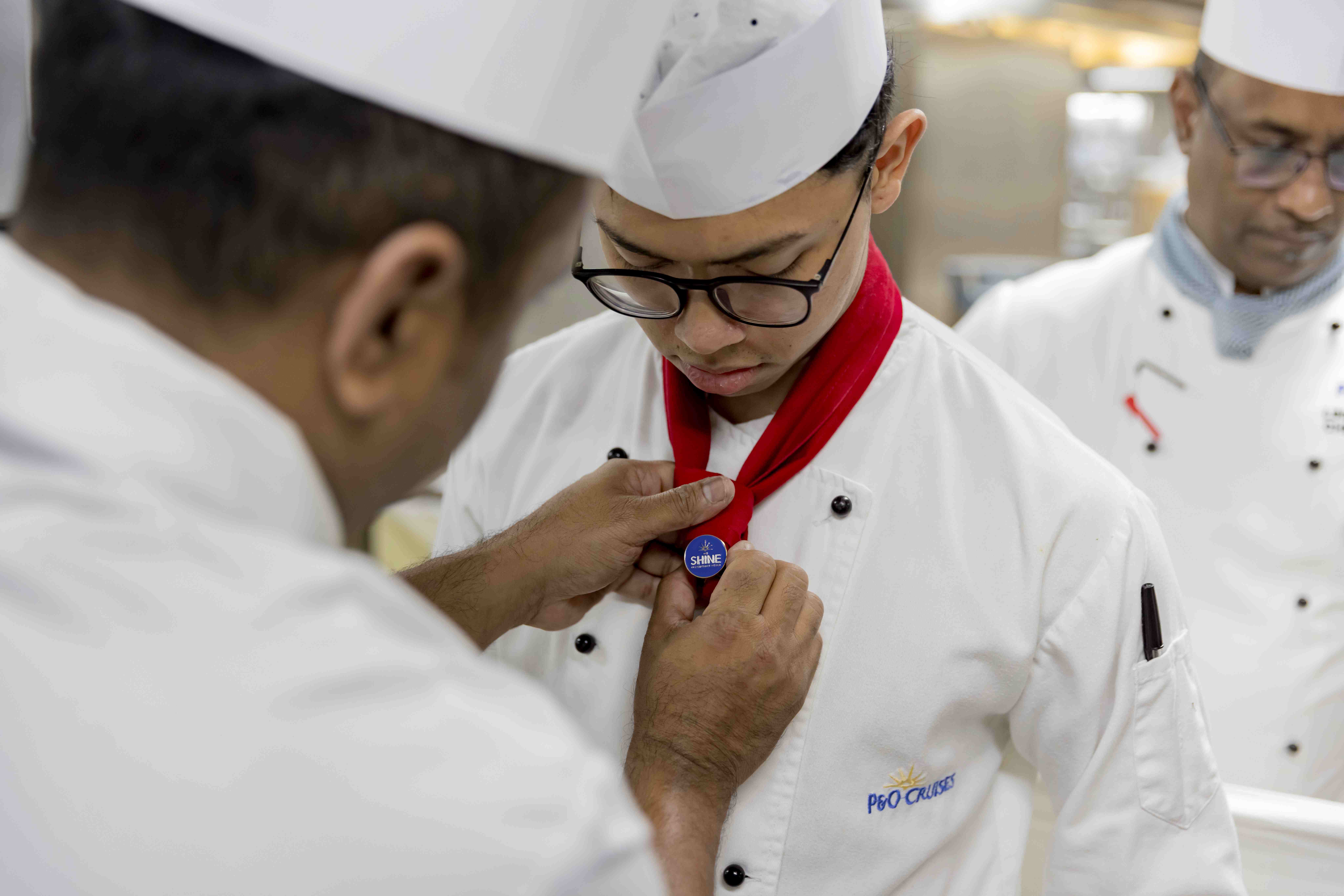 A chef fastens a red neckerchief with a blue badge onto a young chef’s uniform, who wears glasses and a white hat, whilst another chef stands in the background.