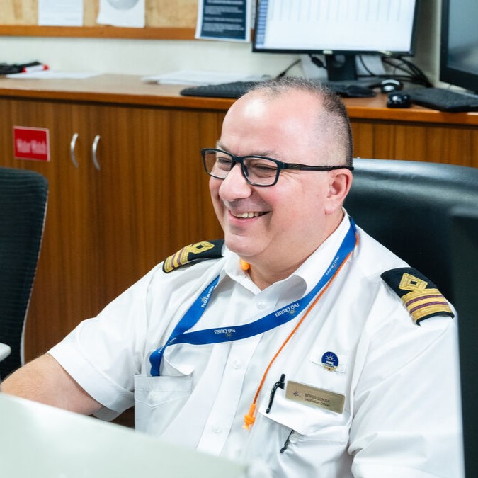 A smiling man in a white uniform with shoulder epaulettes and a name badge sits at a desk in an office, wearing glasses and lanyards—a professional representing P&O Cruises technical careers.