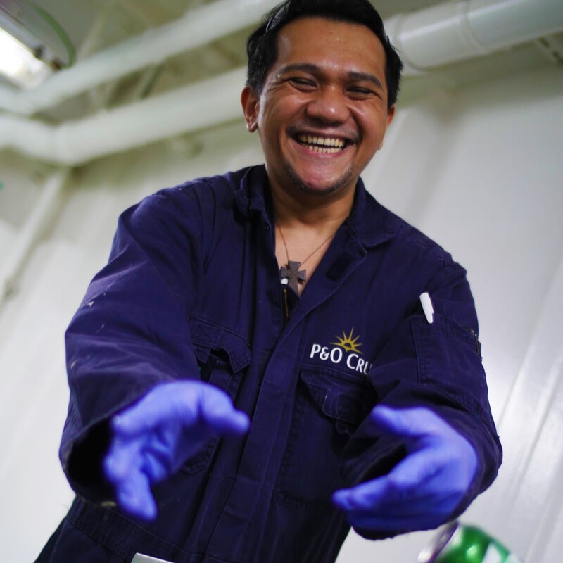 A smiling person in a blue boiler suit and purple gloves reaches forward inside a brightly lit room, showcasing the dynamic world of P&O Cruises technical careers. White pipes and light fittings are visible in the background.