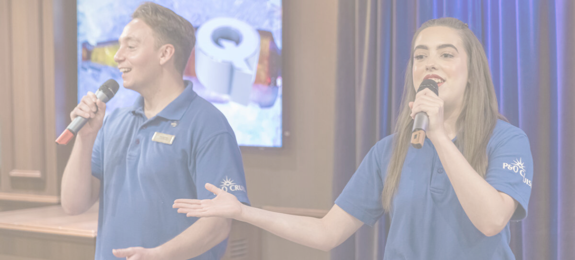 Two people wearing blue polo shirts hold microphones and speak on stage. A screen with colourful visuals is visible in the background, and blue curtains hang to the side.