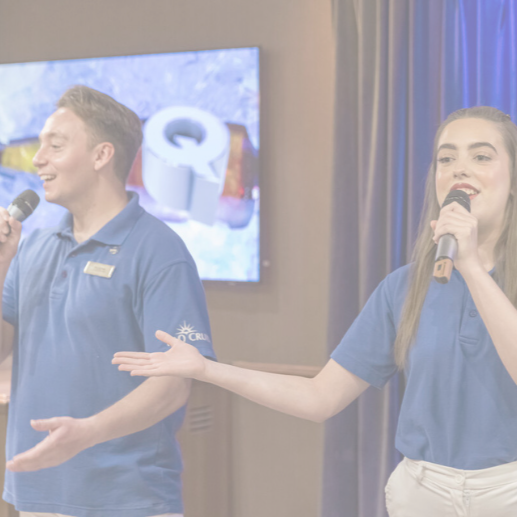 Two people wearing blue collared shirts and holding microphones are speaking on stage. A TV screen with an image and a logo is visible in the background. The scene appears to be indoors.
