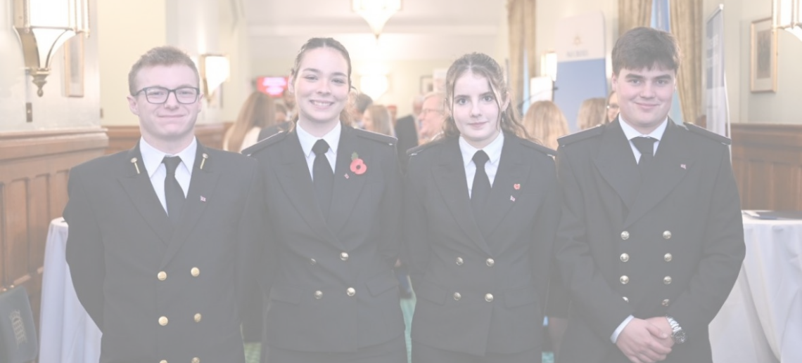 Four young people in formal dark uniforms stand side by side, smiling at the camera in a well-lit room with wood panelling, lamps, and people conversing—showcasing their pride as part of the Carnival Corporation cadet programme.