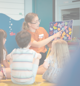 A woman sits on the floor with a group of young children, holding up a colourful board and pointing at it, engaging them in an educational activity.