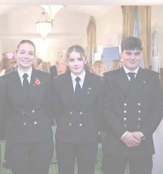 Three young people in dark double-breasted uniforms with gold buttons stand side by side, smiling, in a well-lit room with chandeliers and people in the background, representing the Carnival Corporation cadet programme.