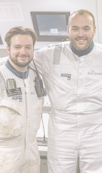Two smiling men wearing white uniforms with P&O Cruises logos stand side by side in a control room, showcasing the teamwork and expertise found in P&O Cruises technical careers. Monitors and equipment fill the background.