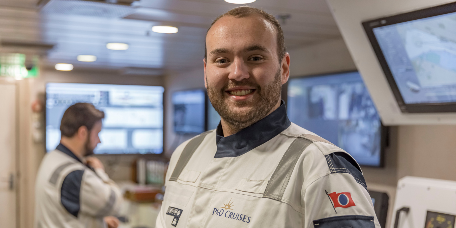 A smiling crew member in a P&O Cruises uniform stands on a ship's bridge, surrounded by control panels and monitors, showcasing the dynamic environment of P&O Cruises technical careers whilst another crew member works behind him.