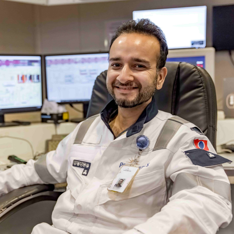 A man in a white uniform with badges and an ID badge sits smiling in an office chair, surrounded by computer monitors and control panels—an inspiring glimpse into P&O Cruises technical careers.