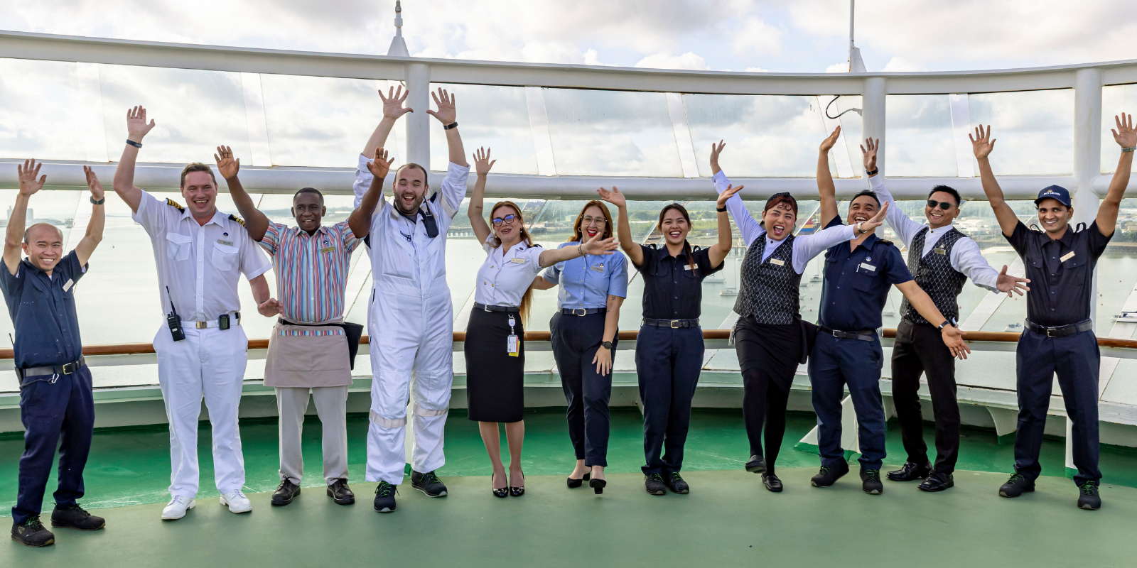 A diverse group of eleven cruise ship crew members in uniform stand in a row on deck, smiling and raising their arms in celebration against a cloudy sky background.