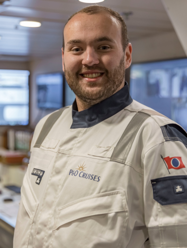 A man wearing a white P&O Cruises uniform with a navy collar and shoulder patches stands indoors, smiling at the camera. The ship's bridge, often the heart of P&O Cruises technical careers, with equipment and windows, is visible in the background.