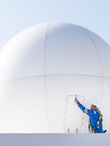 A worker in a blue uniform and safety harness points at a small access door on a large white spherical structure, with a clear sky in the background.